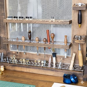 Op de afbeelding: Een houten gereedschapsorganizer met diverse leerbewerkingsgereedschappen. De organizer heeft meerdere planken en doorzichtige acrylhouders voor gereedschappen zoals hamers, ponsen en borstels. De gereedschappen zijn gemaakt van hout en metaal.