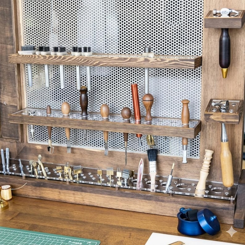 Op de afbeelding: Een houten gereedschapsorganizer met diverse leerbewerkingsgereedschappen. De organizer heeft meerdere planken en doorzichtige acrylhouders voor gereedschappen zoals hamers, ponsen en borstels. De gereedschappen zijn gemaakt van hout en metaal.