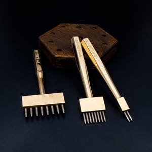 May include: Three gold-colored leather working tools with various prong configurations. The tools have rectangular heads with evenly spaced prongs and long handles. The tools are displayed on a dark surface with a wooden hexagonal base in the background.