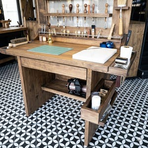 Op de afbeelding: Een houten werkstation voor ambachtslieden in lichtbruin. Het heeft een ingebouwd rek met gereedschap, een groene snijmat en een wit snijplank. Het werkstation heeft een lade en een onderste plank met een machine.