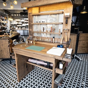 Op de afbeelding: Een houten ambachtelijke werkplek met een ingebouwd rek en opbergruimte. Het bureau heeft een groene snijmat, gereedschap en een rol roze materiaal. De achtergrond toont een winkel met diverse lederwaren.