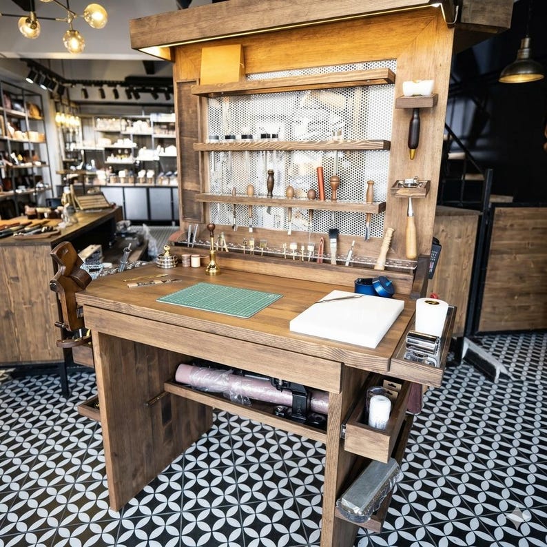 Op de afbeelding: Een houten ambachtelijke werkplek met een ingebouwd rek en opbergruimte. Het bureau heeft een groene snijmat, gereedschap en een rol roze materiaal. De achtergrond toont een winkel met diverse lederwaren.