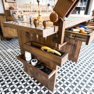 Op de afbeelding: Een handgemaakte houten werkbank met meerdere laden en opbergvakken. De werkbank heeft diverse gereedschappen, waaronder metalen instrumenten, houten handvatten en rollen tape. Het hout heeft een donkere vlek en de vloer heeft een zwart-wit geometrisch patroon.