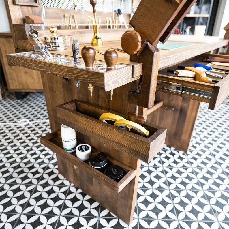 Op de afbeelding: Een handgemaakte houten werkbank met meerdere laden en opbergvakken. De werkbank heeft diverse gereedschappen, waaronder metalen instrumenten, houten handvatten en rollen tape. Het hout heeft een donkere vlek en de vloer heeft een zwart-wit geometrisch patroon.