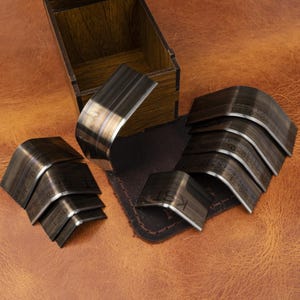 Könnte beinhalten: Ein Satz gebogener Metallwerkzeuge für die Lederverarbeitung mit eingravierten Maßen an den Seiten. Die Werkzeuge sind neben einer Holzkiste und einem dunkelbraunen Lederstück angeordnet, alles auf einer braunen Lederoberfläche.