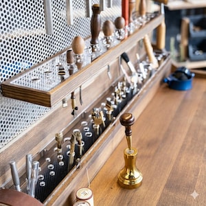 Op de afbeelding: Een houten werkbank met diverse gereedschappen, waaronder hamers, ponsen en polijstmachines, georganiseerd in een trapsgewijs opslagsysteem. De gereedschappen worden tentoongesteld op een houten plank met een geperforeerde metalen achterwand. Een gouden gereedschap staat op de voorgrond.