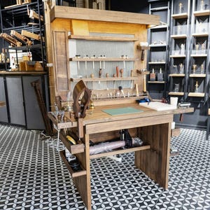 Op de afbeelding: Een houten werkbank met diverse gereedschappen, zoals hamers en priem, voor leerbewerking. De werkbank heeft planken en laden voor opslag, en een groene snijmat op het werkoppervlak. De vloer heeft een zwart-wit geometrisch patroon.