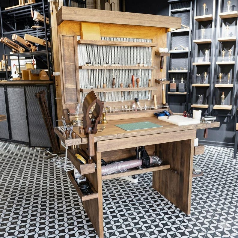 Op de afbeelding: Een houten werkbank met diverse gereedschappen, zoals hamers en priem, voor leerbewerking. De werkbank heeft planken en laden voor opslag, en een groene snijmat op het werkoppervlak. De vloer heeft een zwart-wit geometrisch patroon.