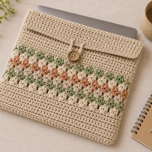 Puede incluir: Funda para portátil beige tejida a ganchillo con cierre de solapa y botón de madera. Presenta un diseño decorativo en verde y melocotón. La funda está diseñada para proteger un portátil, de unos 30 cm de ancho.