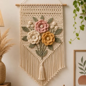 Op de afbeelding: Een macramé wandkleed met een crèmekleurige achtergrond en een bloemenontwerp. De bloemen zijn in tinten roze, crème en geel, met groene bladeren. Het stuk heeft franjes aan de onderkant en is opgehangen aan een houten stok.