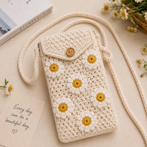 May include: Cream-colored crochet crossbody bag with daisy flower appliques. The bag has a flap closure with a wooden button and a long strap. A small card with the text "Every day can be a beautiful day" is visible.