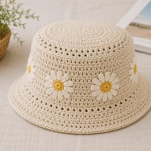 May include: A cream-colored crochet bucket hat adorned with white and yellow daisy appliqués. The hat features a wide brim and a textured, open-weave design, perfect for sun protection. This handmade hat is a stylish accessory for warm weather.