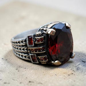 May include: A silver ring with a large, oval-cut, deep red gemstone. The ring has intricate detailing on the band and around the gemstone setting, with smaller red stones accenting the design. The ring is displayed on a textured surface.