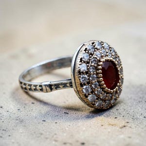 Könnte beinhalten: Silberring mit einem ovalen, roten Edelstein in der Mitte, umgeben von kleinen, klaren Steinen. Das Ringband hat ein dekoratives Muster. Der Ring liegt auf einer strukturierten, hellgrauen Oberfläche.