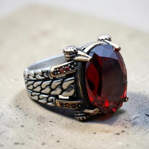 Puede incluir: Un anillo de plata con una gran piedra preciosa ovalada de color rojo intenso. El anillo tiene una banda detallada y texturizada y detalles en metal dorado. La piedra preciosa es el punto focal, añadiendo un toque de lujo.