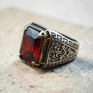 May include: A silver-toned metal ring featuring a large, rectangular, deep red faceted gemstone set in the center. The band is decorated with intricate engraved scrollwork patterns and a geometric Greek key border along the sides of the setting.