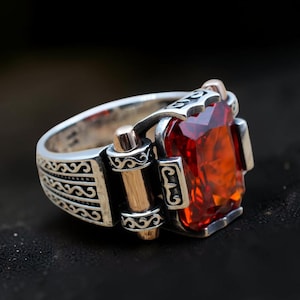 May include: A silver ring with a large, rectangular, orange-red gemstone. The ring has intricate detailing on the band and gold accents. The gemstone is the focal point, catching the light and adding a touch of elegance.