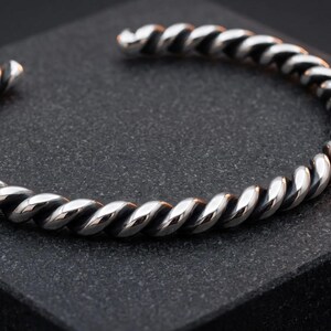 Puede incluir: Un brazalete tipo brazalete de color plateado con un diseño de cuerda trenzada continua. La superficie metálica tiene un acabado pulido en las secciones elevadas y un aspecto oscuro y oxidado en las ranuras del patrón en espiral.