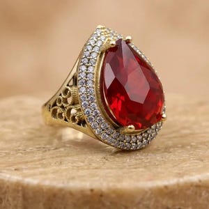Könnte beinhalten: Ein goldfarbener Ring mit einem großen, birnenförmigen, tiefroten facettierten Edelstein in der Mitte. Der zentrale Stein ist von einem Heiligenschein aus kleinen, klaren, rund geschliffenen weißen Steinen umgeben. Die Ringschiene zeigt filigrane Durchbrucharbeiten.