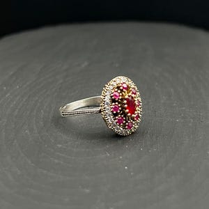 May include: An oval-shaped ring featuring a central red stone surrounded by a cluster of smaller red stones and a border of tiny white stones. The setting is gold-toned with a textured silver-toned band that has a beaded edge detail.