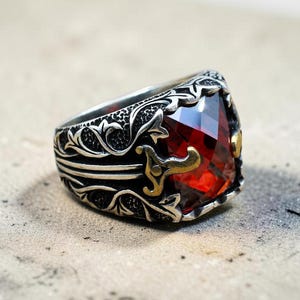 Op de afbeelding: Een zilverkleurige ring met een gedetailleerd, donker geoxideerd bloemmotief in reliëf op de band. Het middelpunt is een grote, gefacetteerde, dieprode edelsteen, geaccentueerd door kleine, gepolijste goudkleurige metalen details naast de zetting.