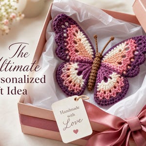Op de afbeelding: Een gehaakte vlinder in tinten paars, roze en crème met een bruin lichaam, rustend in een roze geschenkdoos bekleed met wit vloeipapier. Een label aan de vlinder bevat de tekst "Handmade with Love" met een hartje. De tekst luidt "The Ultimate Personalized Gift Idea."