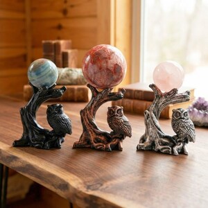 May include: Three decorative stands shaped like tree branches, each featuring a perched owl figure at the base. The stands hold spherical stones: a blue and white striped sphere, a red and white marbled sphere, and a pale pink translucent sphere.