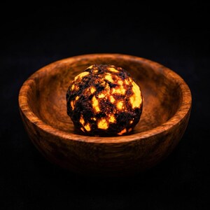 May include: A glowing, textured sphere with a dark, charred exterior and bright orange light emanating from within, resting inside a smooth, polished wooden bowl with a visible grain pattern against a solid black background.
