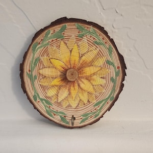 May include: A round, wooden wall hanging featuring a hand-painted sunflower design. The sunflower is yellow and orange, with a brown center. A green and red leaf border surrounds the flower. A small metal hook is attached to the bottom.