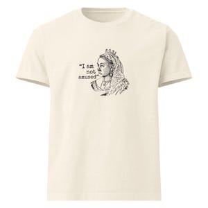 May include: Cream-colored t-shirt featuring a black and white illustration of Queen Victoria with the text "I am not amused." The graphic is on the front of the shirt. The shirt has short sleeves and a crew neck.