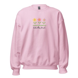 May include: A pale pink sweatshirt with three embroidered flowers in yellow, pink, and lilac, with green stems and leaves. The words "totally feral" are written in a script font below the flowers. The sweatshirt has a crew neck and long sleeves.