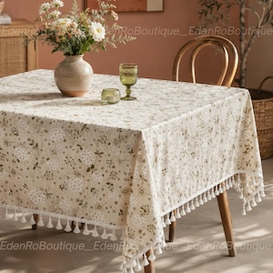 May include: A square tablecloth with a beige background featuring a repeating pattern of small white and brown floral motifs. The edges of the fabric are finished with a decorative white fringe trim. A ceramic vase with flowers sits on top.