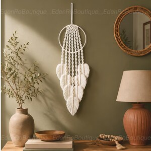 Puede incluir: Un colgante de pared de macramé blanco con una parte superior circular y detalles en forma de pluma. La decoración de estilo bohemio se exhibe contra una pared verde apagada, con un espejo redondo y una lámpara en el fondo.