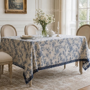 Puede incluir: Un mantel rectangular con un estampado floral azul y blanco tipo toile, con un borde de encaje festoneado en azul oscuro. La tela cubre una mesa de comedor, decorada con un jarrón de cristal con flores blancas y un cuenco blanco con tapa.