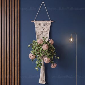 May include: A cream-colored macrame wall hanging featuring a butterfly pattern and a central pocket holding a green plant with round, light pink flower clusters. The piece is suspended from a wooden dowel with a cord and ends in a long fringe.