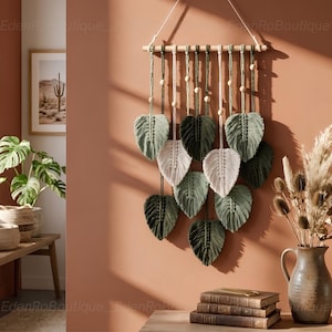 May include: A macramé wall hanging with leaf-shaped designs in shades of green and white. The leaves are suspended from a wooden dowel with wooden beads. The wall decor is displayed against a peach-coloured wall.