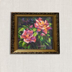 May include: An oil painting of two roses in full bloom, with pink and yellow petals, surrounded by green leaves. The artwork is in a gold-colored ornate frame. The artist's signature, LLAURADO, is visible in the lower right corner.
