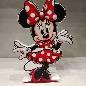 Può includere: Una figura sagomata autoportante di Minnie Mouse con il suo iconico vestito rosso a pois bianchi, un fiocco rosso abbinato, guanti bianchi e scarpe rosse. Il personaggio è raffigurato in una posa allegra, montato su una base bianca.