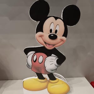 Peut inclure: Une figurine plate découpée représentant Mickey Mouse avec ses oreilles noires emblématiques, un visage souriant, un torse noir, des gants blancs, un short rouge avec deux boutons ovales blancs et de grandes chaussures jaunes.