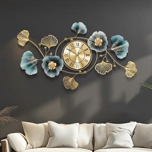 May include: A decorative wall clock featuring a gold-toned clock face with Roman numerals, surrounded by a floral design. The design includes gold-colored leaves and blue and gold flowers. The clock is mounted on a dark gray wall.