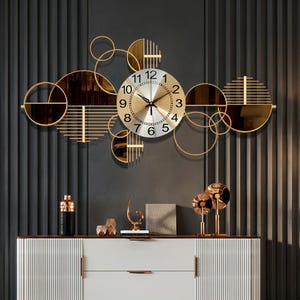 May include: A gold-toned wall clock with a circular face and Roman numerals. The clock is surrounded by a decorative arrangement of gold circles, mirrored elements, and geometric shapes. The clock is mounted on a dark gray wall above a white cabinet.