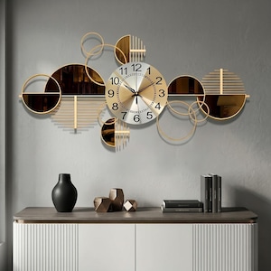 May include: A gold and silver wall clock with a circular face and black numerals. The clock is surrounded by gold geometric shapes, including circles and semi-circles. The clock is mounted on a gray wall above a white cabinet.