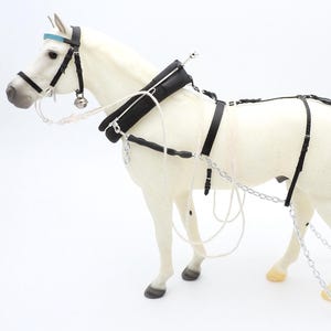 Puede incluir: Una figura de caballo blanco con un arnés de cuero negro que incluye una testera azul claro, un cascabel plateado y cadenas de metal plateado. El arnés cuenta con un collar acolchado, riendas y correas que llegan a una barra de madera.