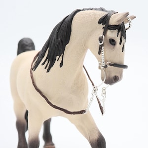 Puede incluir: Una figura de caballo blanco con una crin y cola negras, que lleva un bozal de cuero marrón y una boquilla de plata. El caballo está de pie sobre un fondo blanco.