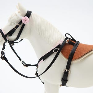 K&ouml;nnte beinhalten: Ein Miniatur-Pferdesattel- und Zaumzeug-Set. Das schwarze Zaumzeug hat rosa Akzente und eine kleine rosa Blume. Der Sattel ist braun mit einer cremefarbenen Unterseite. Das Set ist f&uuml;r ein Spielzeugpferd konzipiert.