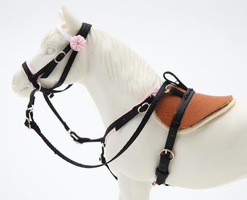 K&ouml;nnte beinhalten: Ein Miniatur-Pferdesattel- und Zaumzeug-Set. Das schwarze Zaumzeug hat rosa Akzente und eine kleine rosa Blume. Der Sattel ist braun mit einer cremefarbenen Unterseite. Das Set ist f&uuml;r ein Spielzeugpferd konzipiert.
