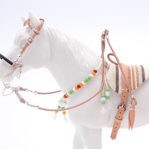 Puede incluir: Una figurita de caballo miniatura blanca que lleva una brida de cuero marrón, una silla de montar y un collar de cuentas. La silla de montar es de rayas marrones y blancas.