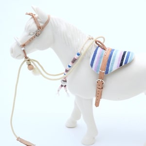 Puede incluir: Un caballo de juguete blanco con una silla de montar y brida en miniatura. La silla tiene rayas azules, rosas y blancas. La brida y las correas de la silla son de cuero color canela. Las riendas son una cuerda color crema con cuentas decorativas.