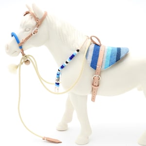 Puede incluir: Un caballo de juguete blanco con una montura y brida en miniatura. La montura es de rayas azules y grises, y la brida es de color beige con detalles azules. Las riendas y otros accesorios son de color beige y marrón.