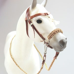 Puede incluir: Un modelo de caballo blanco con una cabezada de cuero marrón con un bozal de cuerda trenzada y una boquilla de plata.